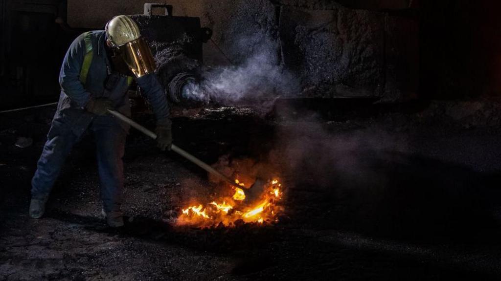 A worker dressed in a blue overall and white helmet raking hot coal in blast furnace number four at Tata's Port Talbot steelworks.