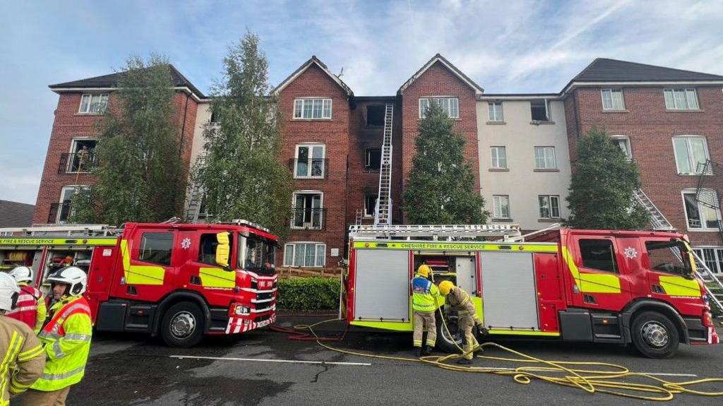 Fire engines and firefighters work outside a block of flats which has been blacked by fire damage
