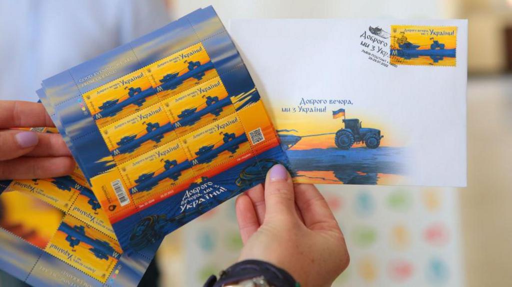 Hand holds stamps that read "Good evening, we're from Ukraine", showing a Ukrainian tractor pulling along a Russian tank