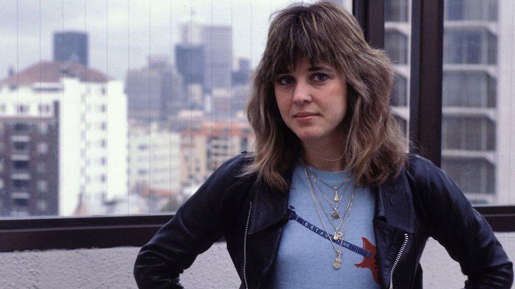 Suzi Quatro wearing a blue top and black jacket while standing on a rooftop. Behind her is a window, which has the reflection of buildings in it.