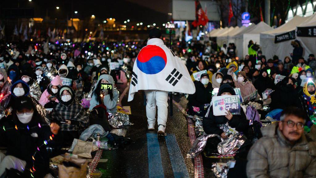 A man wearing a South Korean flag as a cape walks through a sea of anti-Yoon protesters sitting on the ground.