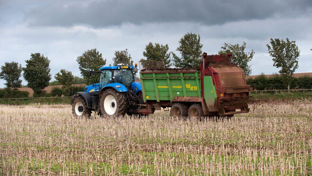 A blue tractor pulling a green trailer spreading fertiliser in a stubble field