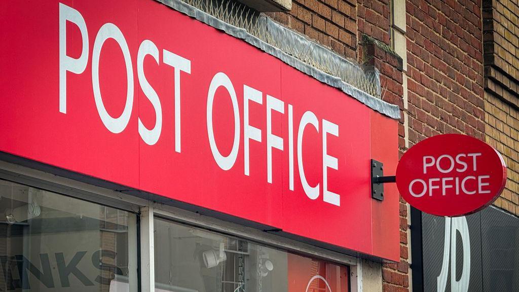 The outside of a Post Office branh in Taunton, England in February last year. There is a big red Post Office sign, with a JD Sports store next door.