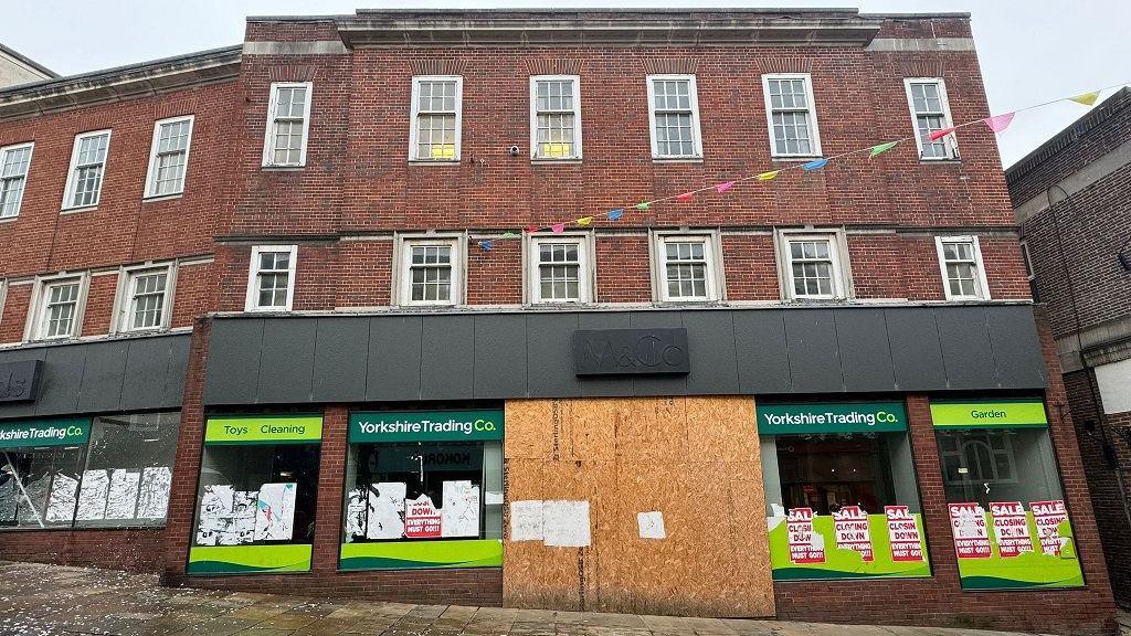 A three-storey brick property. Above the ground-floor windows are green signs with white lettering for the Yorkshire Trading Company. The doors of the building are boarded up and several tatty and partly removed red-and-white "Closing down sale" posters are in the windows.