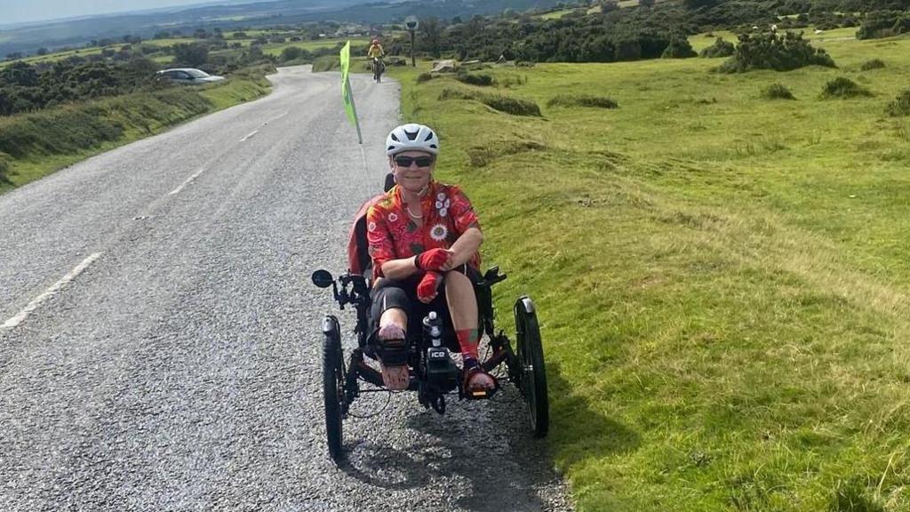 A woman sitting on a trike on a hilly road on a moor top. She is wearing a red cycling top and a white helmet. To the right is a grassy field. Behind her are trees and shrubs. In the background a cyclist can be seen climbing up the road.