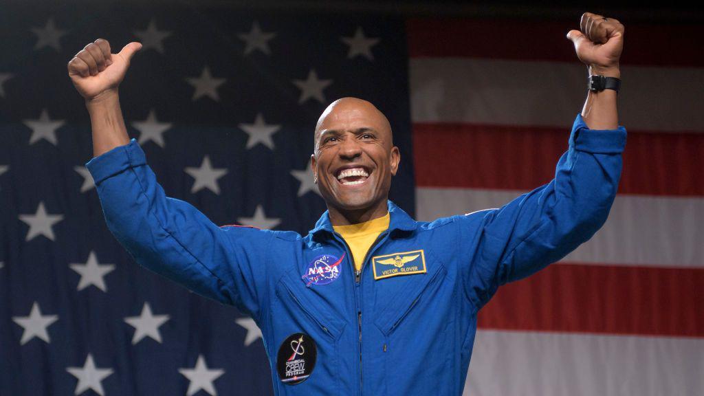victor glover in a blue jacket smiling and holding both thumbs up in front of an American flag