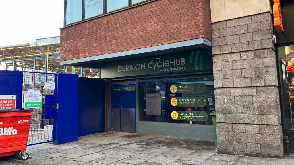 The outside of Derbion Cycle Hub. It has a blue double door on the left, window on the right and a sign above on a green background which reads "cyclehub". On the left, is building work where the the former Eagle Market was. 