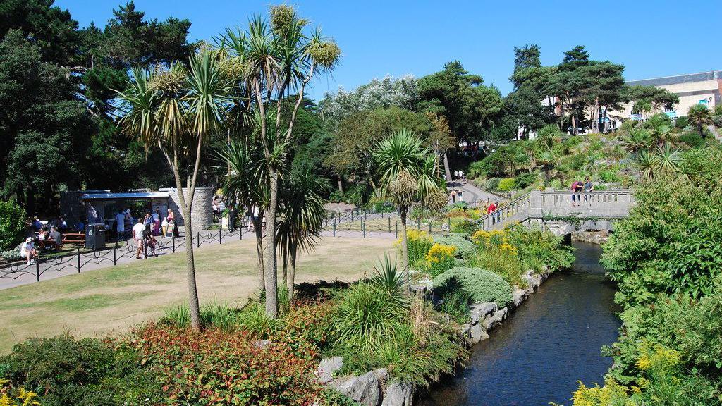 Bournemouth's Lower Gardens - several palm trees and the River Bourne can be seen in the foreground with people walking along the pathways in the background