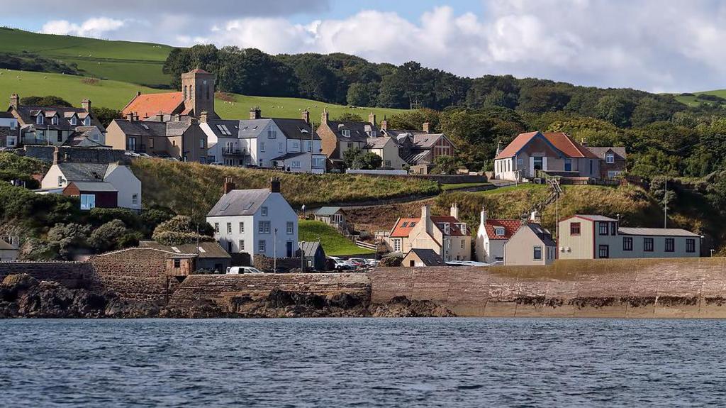 A small village of grey and red-roofed houses above a harbour, with the sea in the foreground