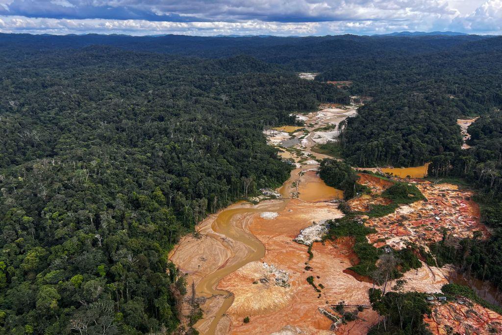 A stretch of green Amazon rainforest stretches to a faraway horizon of blue mountains and sky but in the foreground a wide stretch of land around a river is laid bare and brown