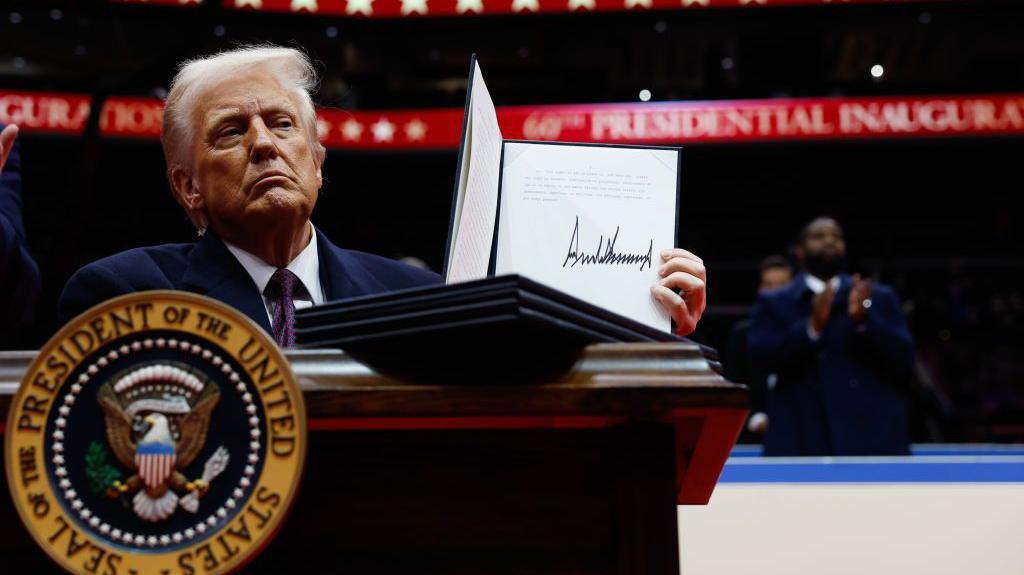 President Donald Trump holds up an executive order after signing it during an indoor inauguration parade at Capital One Arena on January 20. He stands before a podium with a presidential seal. HIs expression is neutral. 