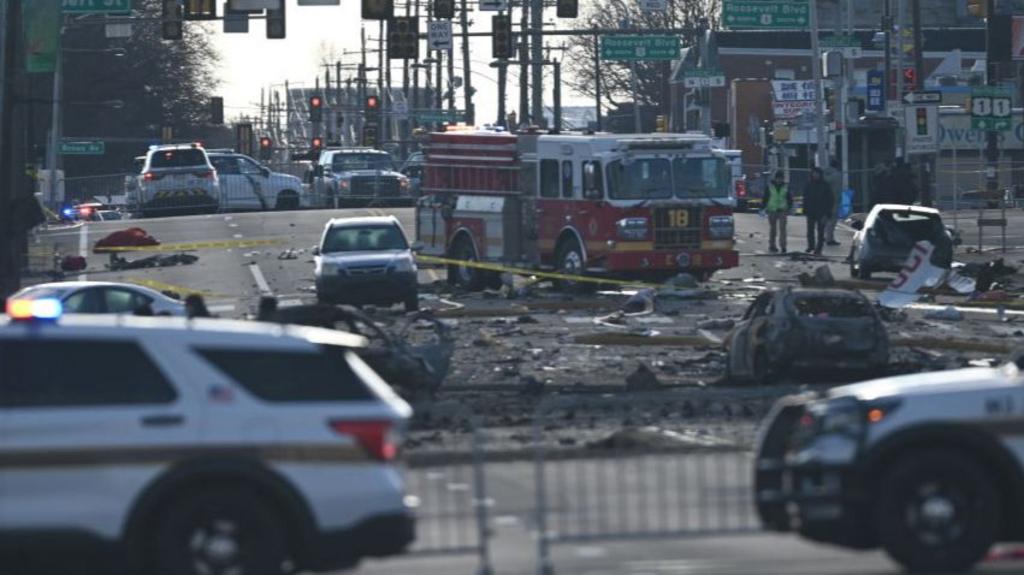 Fire engine parked in the middle of the street surrounded by charred debris of cars and crashed plane. In the foreground, blurred, are two white police cars