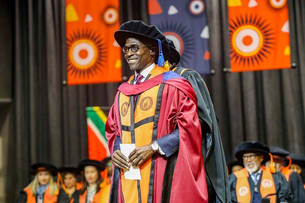 A man wearing academic robes stands on stage and smiles.