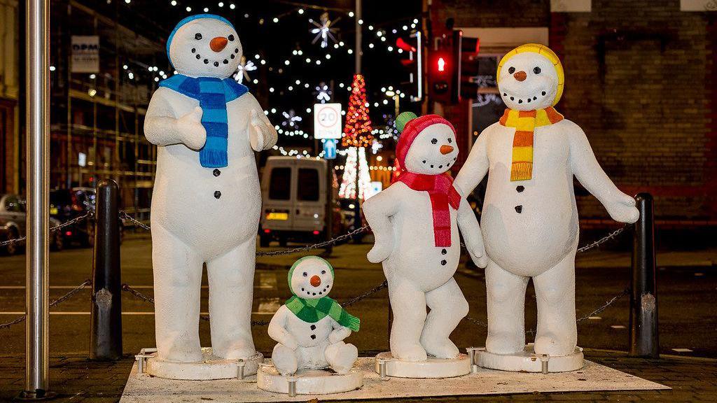 Four plastic snowmen pose in the street, two look like children snowmen, with a Christmas tree and Christmas lights in the background. 