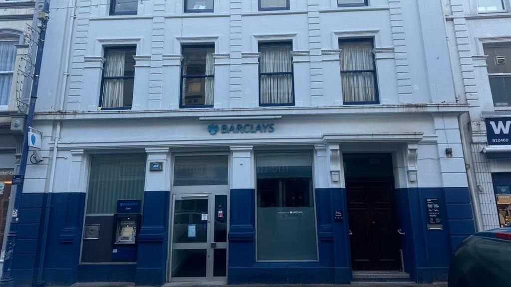 A wider look at  the exterior of the Barclays branch in Ramsey featuring a blue sign with the name of the bank. The walls are painted blue and white, and there is a brown wooden door, clear double doors, and features a cash point machine.