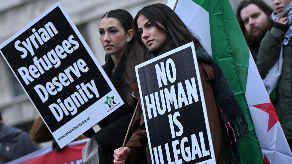 Two women stand outside 10 Downing Street holding Syrian flag and placards reading: 'Syrian refugees deserve dignity' and 'no human is illegal'.