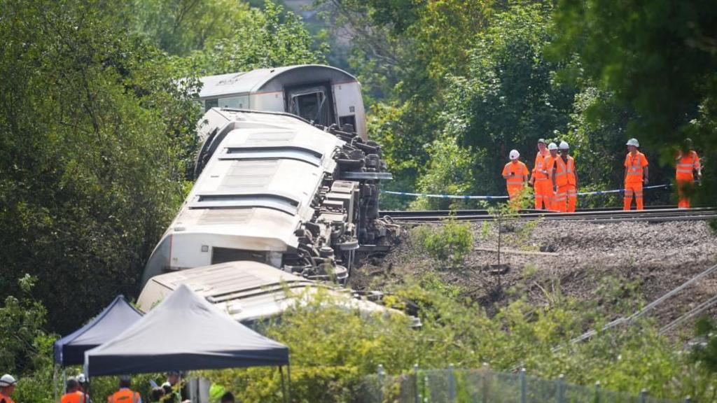 Three train carriages on their side while emergency workers stand watching