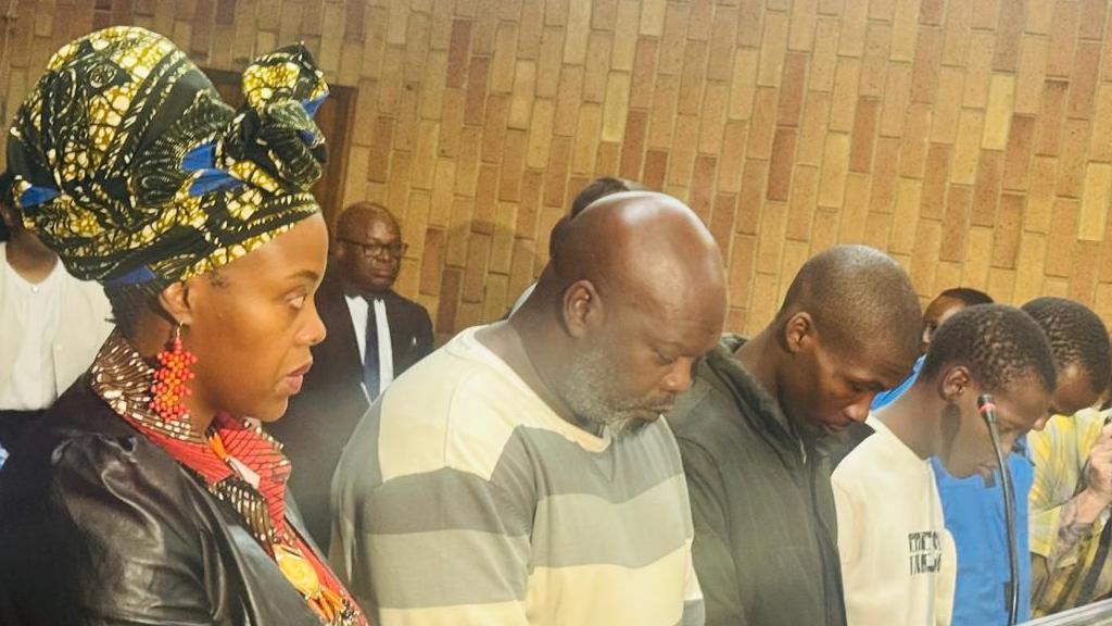 Nonkululeko Patricia Mantula, wearing a headwrap, large beaded orange, red earrings and a leather jacket look on as she sits at the end of a row in court next to her four other accused - all men - who have their heads bowed.