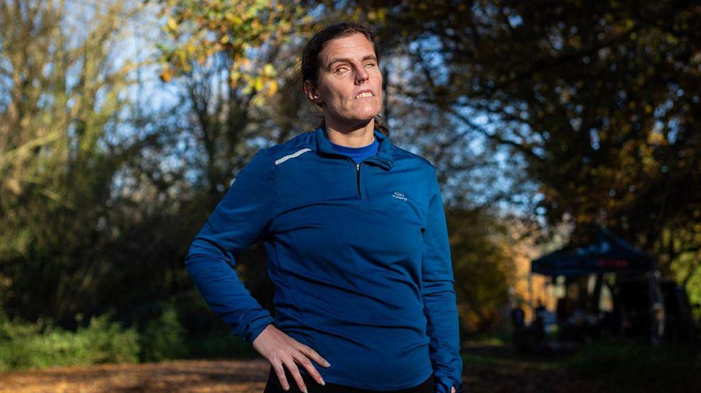 A woman with tied-back brown hair, wearing a navy blue running top and dark bottoms. She is stood with one hand on her hip in a forest setting with lots of trees behind her.