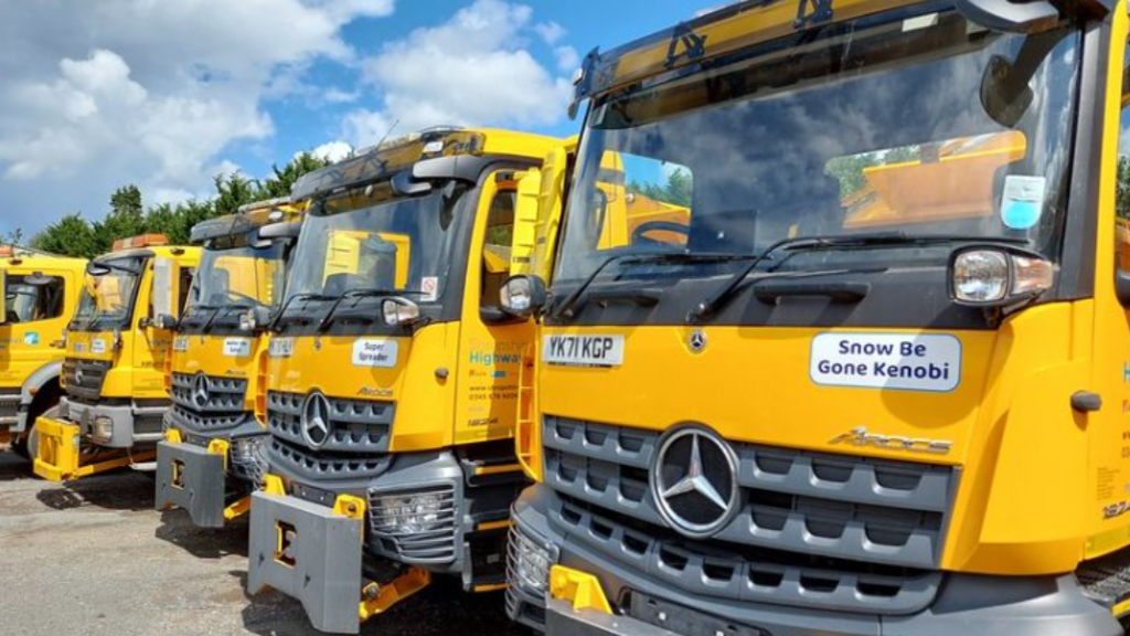 A line of five yellow gritter lorries, the closest to the camera has a sticker proclaiming 'Snow be Gone Kenobi'