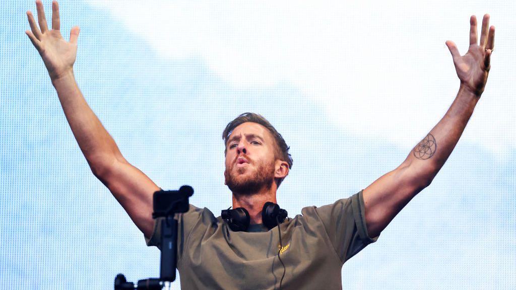 Calvin Harris stands behind a mixing desk with headphones around his neck. He has his arms raised in the air