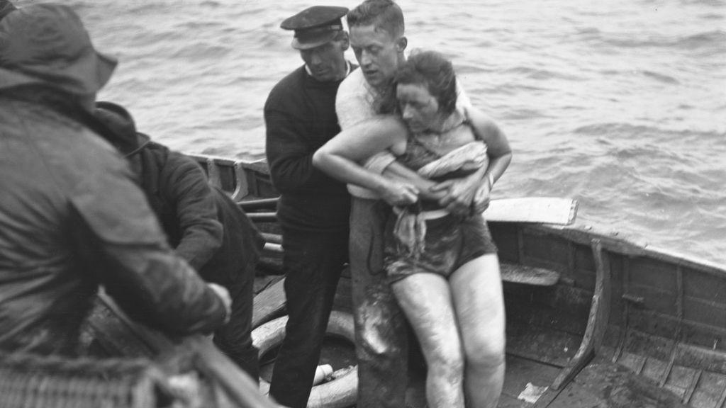 Two men on a wooden row boat at sea holding onto a woman in a swimming costume, who looks exhausted. In the black and white image from 1927, they are preparing to hand her over to people in a larger boat.