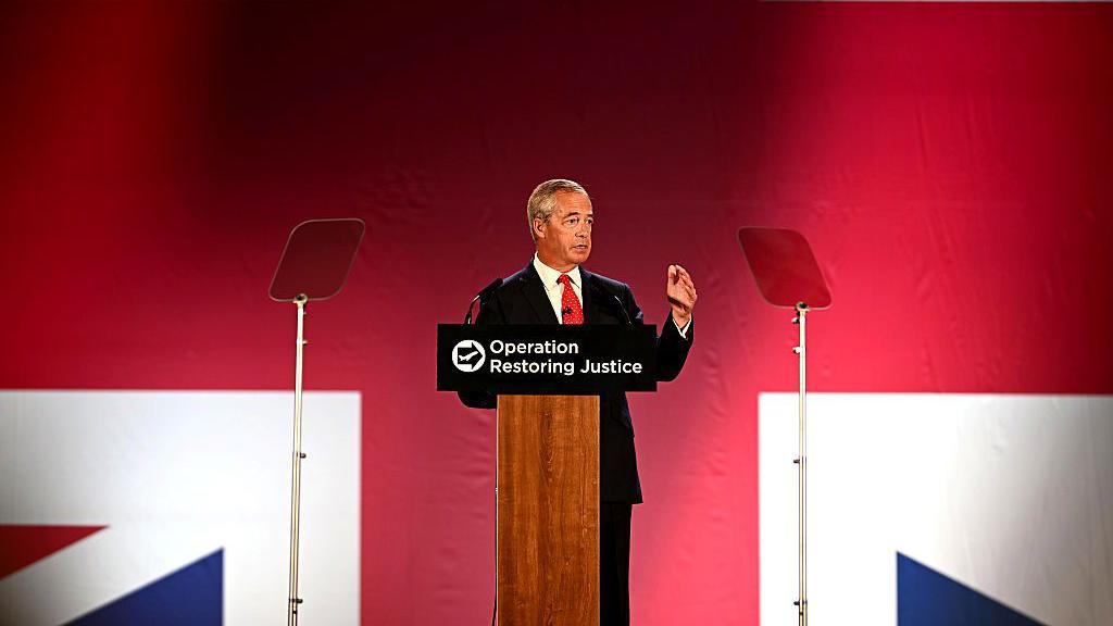 Nigel Farage, wearing a black suit and a red tie, standing at a podium that says "Operation Restoring Justice" on it. He's talking into a microphone, with a large Union Jack projected onto the wall behind him.