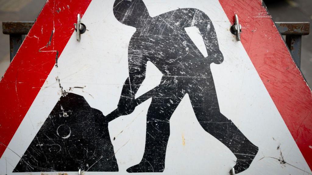 A close up image of a road work sign, where a blacked out man shape is shoving roads. It has a red boarder and a white background. 