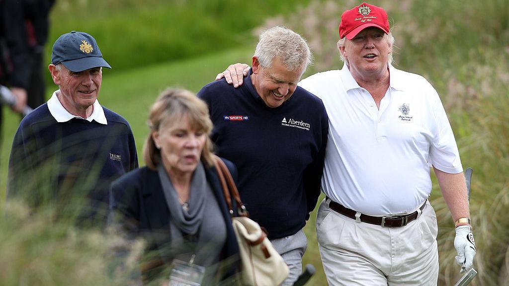 Colin Montgomerie, who has short, white hair, is wearing a dark blue branded crew-neck jumper and light chinos. He is walking along, smiling. Donald Trump, in white polo shirt, light chinos and red baseball cap, is walking alongside him, his right arm draped over Monty's shoulder. A woman in business dress and a man in blue jumper and white polo shirt walk beside them. They are on a golf course. In the rough, by the look of things.