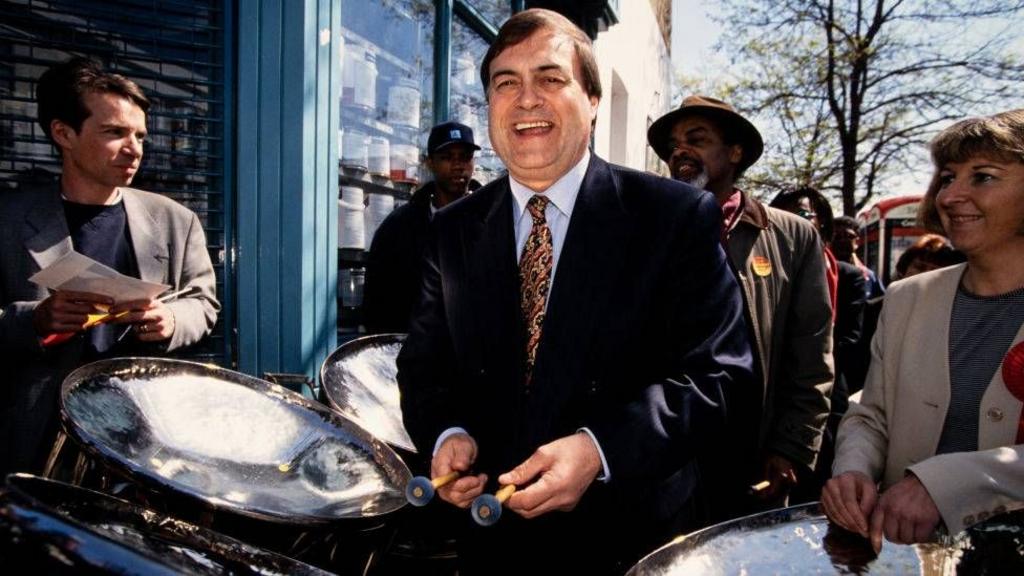 John Prescott is seen campaigning alongside supporters. There are brass drums surrounding him and he has a big smile on his face.