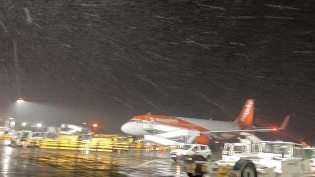 An easyJet plane at Bristol airport, at night, with snow falling. There are white trucks close by with yellow barriers in the background. 