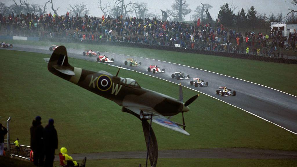 The Williams of Alain Prost leads on the first lap of the wet 1993 European Grand Prix at Donington Park with team-mate Damon Hill behind, followed by Karl Wendlinger's Sauber and the McLaren of Ayrton Senna. A model of a Spitfire aircraft can be seen in the foreground