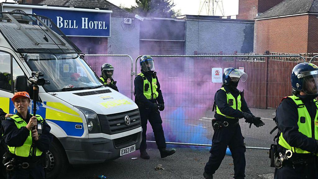 Police respond to a protest outside the Bell Hotel in late July. The hotel's entrance can be seen behind a tall, temporary metal fence, outside of which a police van is parked and five uniformed officers are emerging. Red and blue smoke is visible near the hotel's entrance, from flares lit by protesters.