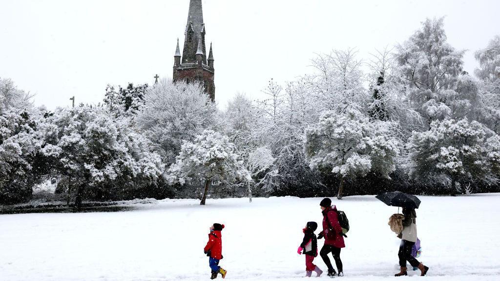Snowy scene with a park area and a church in the background.  Trees covered in snow and a group of people walking in the snow.