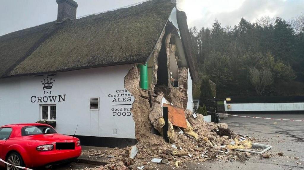 The pub in the aftermath of the collapse. The thatched two-storey building is covered in white render. The end of the building beneath the chimney is missing and the inside is visible. Huge piles of rubble are strewn on the ground.