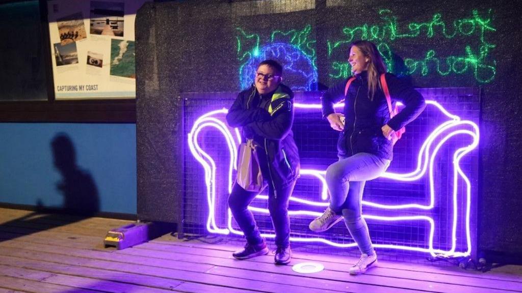 Two people pretend to sit on a sofa created with lights.