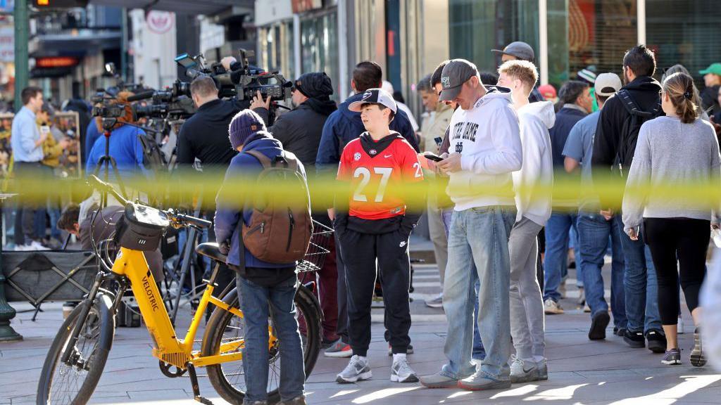 Crowds of people gather on a New Orleans street as police tape can be seen in a blur in the foreground