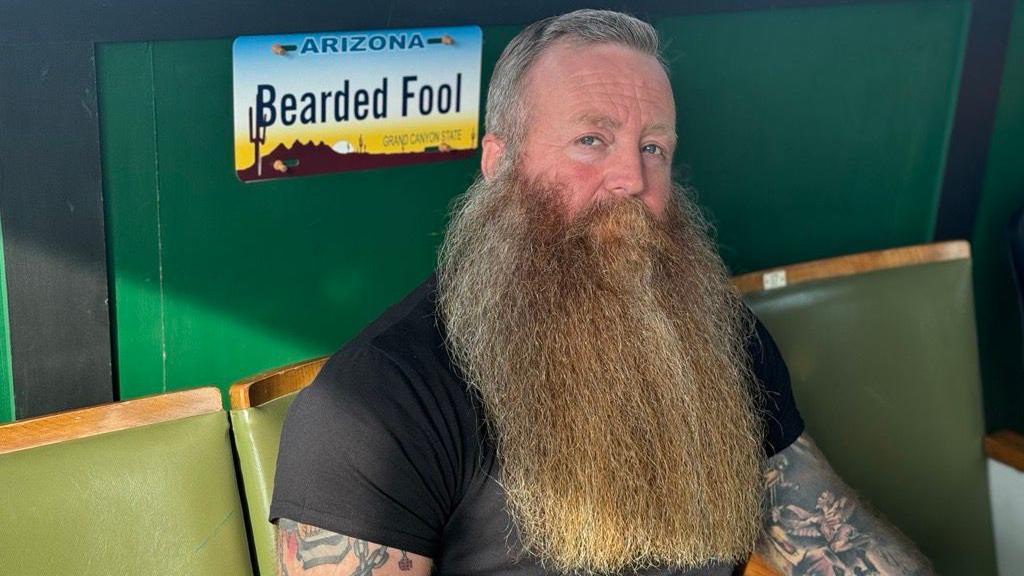 Brian has short grey hair and a long beard and is wearing a black t-shirt and arm tattoos.