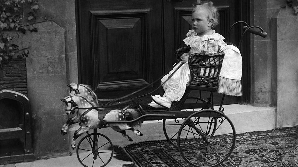 Prince Albert sits in the galloping gig in an old black and white photo. He is holding the reins and wears a traditional white Victorian garment for children. The gig is like an old pushchair with a seat and in front there are two small wooden horses which move when pushed. The picture has been taken in front of a large set of doors.