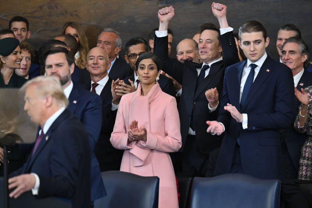 A crowd of people in dark suits surround Donald Trump, including Elon Musk, who has his fists raised in jubilation 