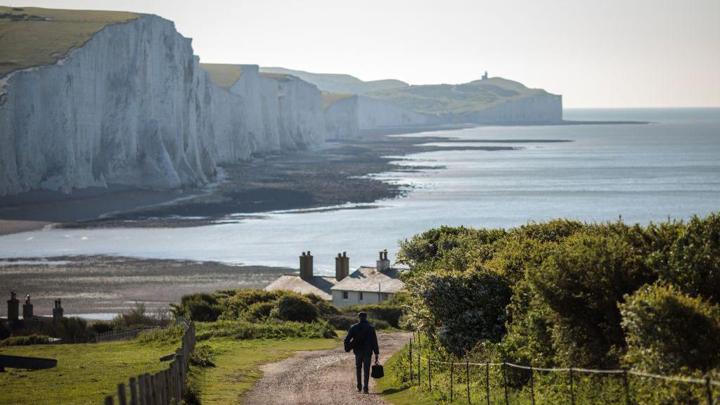 A man walking towards the mouth of the Cuckmere River, with the Seven Sisters cliffs in the background.