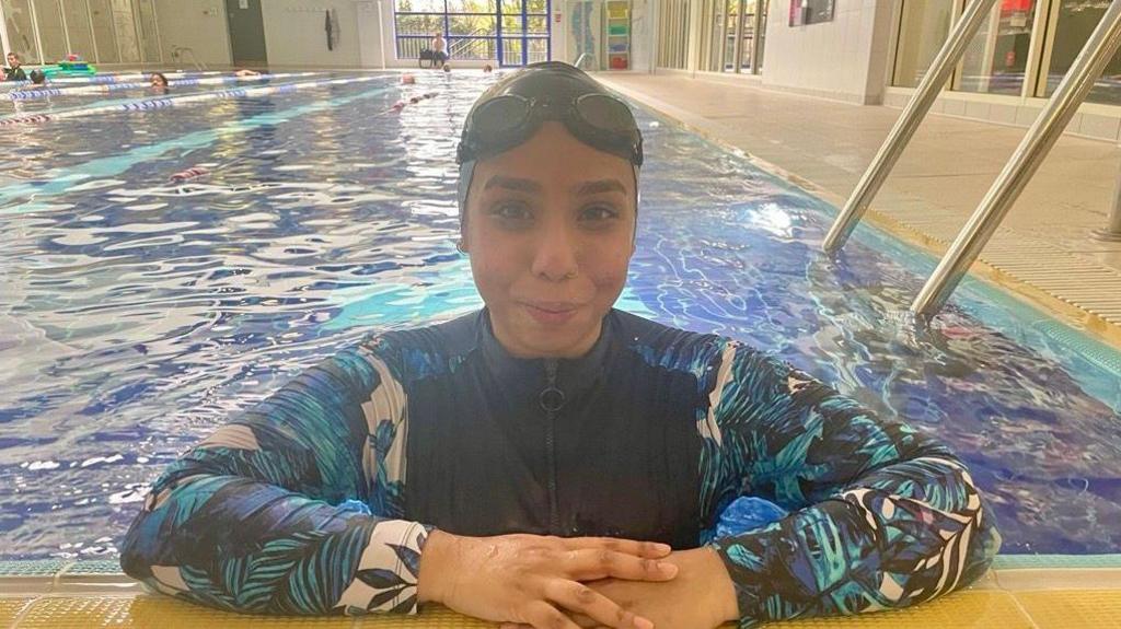 Parin Begum is in the pool and is stood at the edge with her arms resting on the side. She wears a swim cap and goggles and a blue white and black burkini which has patterns of leaves on the sleeves. She is smiling at the camera after finishing the swim challenge.