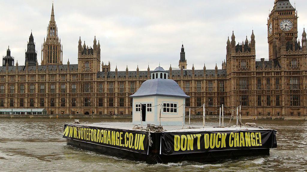 A mock giant duckhouse on a floating platform is towed along the river Thames, with the Houses of Parliament visible in the background. There are black banners with writing in yellow capital font on the sides of the platform, one listing the campaign group website www.voteforachange.co.uk and another with the slogan "Don't Duck Change!".