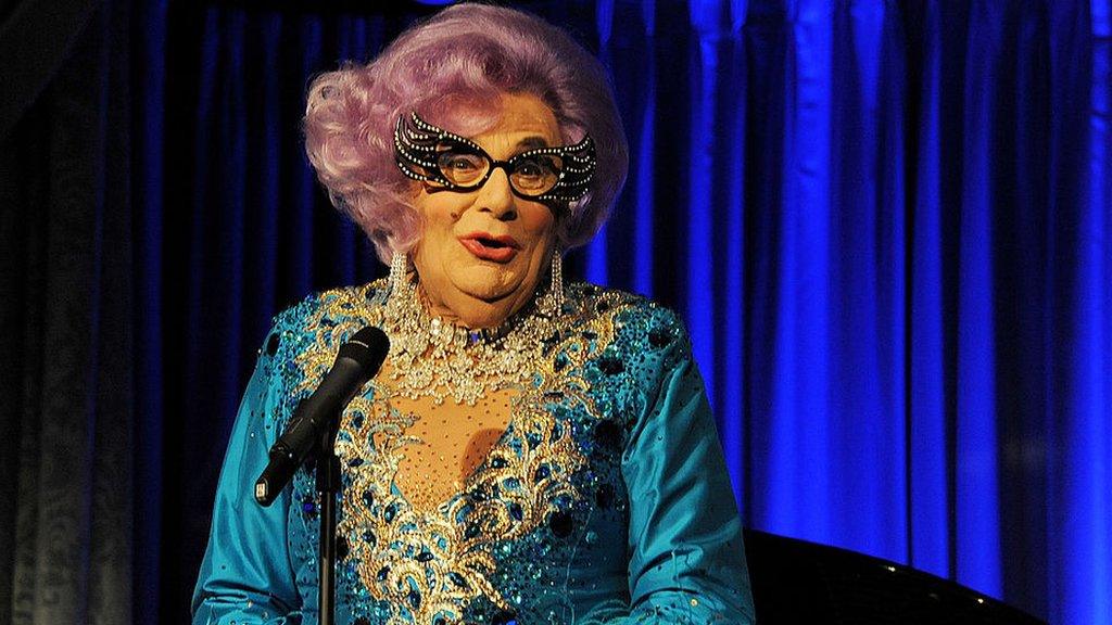 An elderly drag queen with purple hair, large glasses and a blue dress on a stage