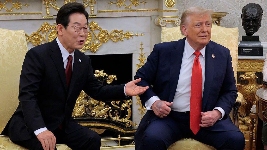 Lee Jae Myung and Trump sitting side by side in yellow armchairs in the Oval Office. They are wearing dark coloured suits with red ties. They're looking and smiling in the same direction.