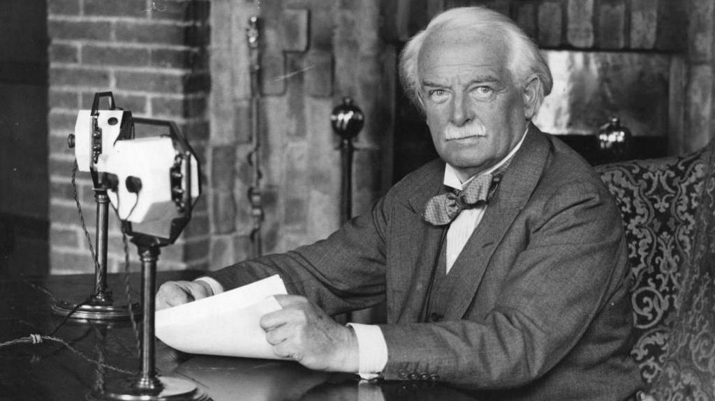 Llun o David Lloyd George yn eistedd wrth ymyl ddesg gyda meicroffon o'i flaen. Mae'n gwisgo siwt ac yn dal papurau yn ei law.
