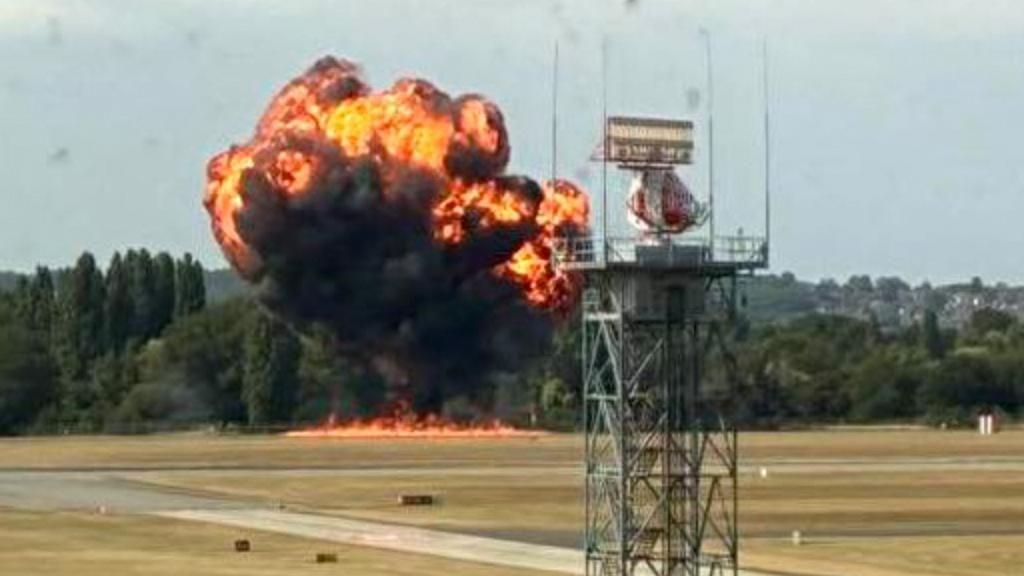 A large ball of flames rises from an airport's runway area. There are flames at its base, with black smoke rising and orange/yellow flames visible within the smoke. An airport communication tower/mast is visibile in the foreground. There is woodland on the far edge of the airport.