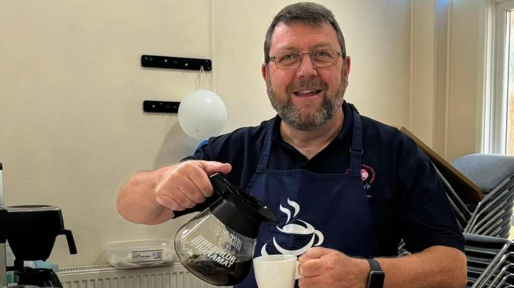 Chris Harper with a beard and glass, wearing a navy T-shirt and navy apron. He is pouring coffee from a jug into a white mug while smiling into the camera.