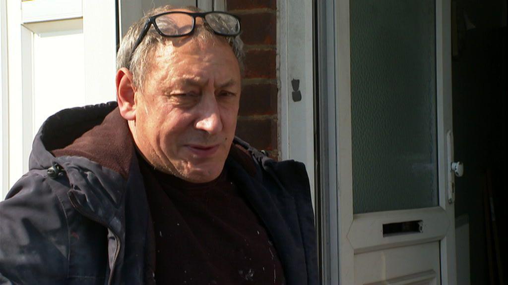 A man with grey hair and glasses on his head is standing outside a house with the door open. He is wearing a large black coat with a brown jumper
