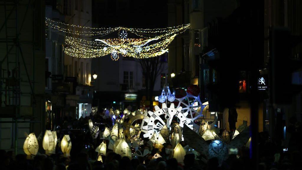 Dozens of people holding up lanterns. There are buildings either side of the street. 